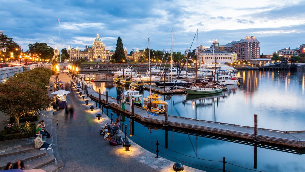 Haven van Victoria bij avondlicht met verlichte gebouwen, boten en mensen op de promenade – reizen met Explore