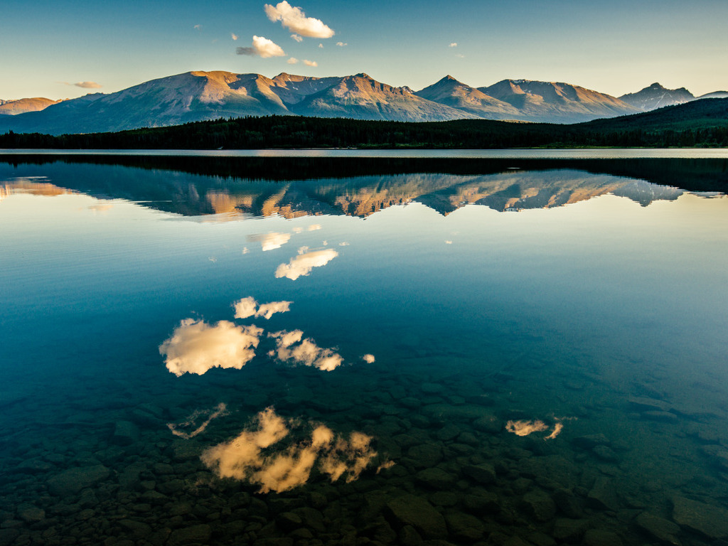 Reflectie van bergen en wolken in het stille water van een meer, omringd door een bos en een blauwe lucht – Canada – reizen met Explore.