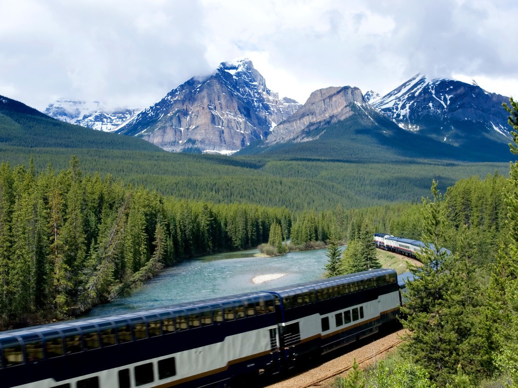 Rocky Mountaineer langs een rivier in de bergen, omringd door dichte bossen en besneeuwde toppen – Canada – reizen met Explore.