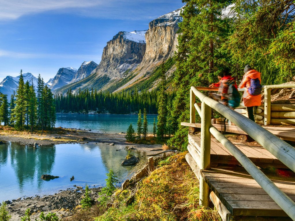 Uitzicht op Spirit Island in Maligne Lake met omringende bergen en een houten platform met hikers – Canada – reizen met Explore.