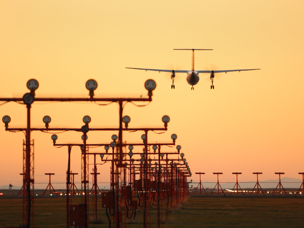 Een vliegtuig nadert de landingsbaan in de avondschemering, begeleid door een rij lampen op hoge palen die het pad naar de luchthaven markeren – reizen met Explore.
