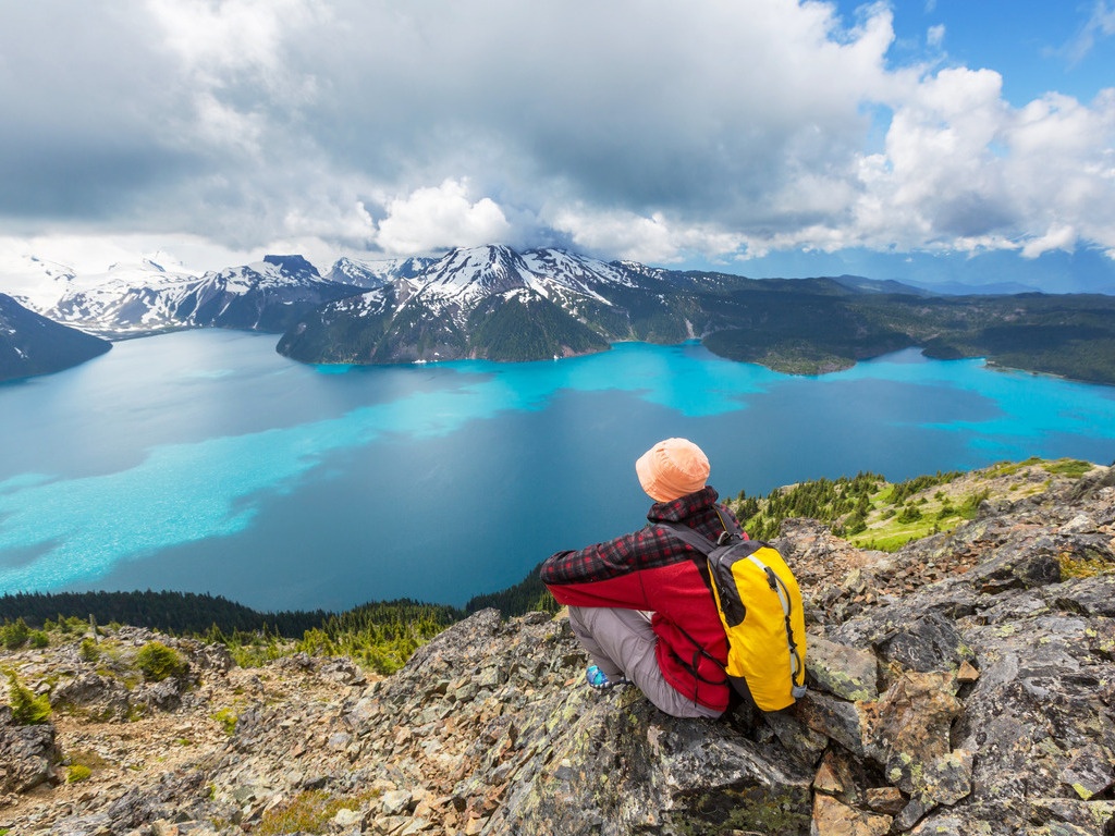 Wandelaar met gele rugzak die uitkijkt over een helder blauw bergmeer omgeven door besneeuwde bergen, Canada – reizen met Explore.